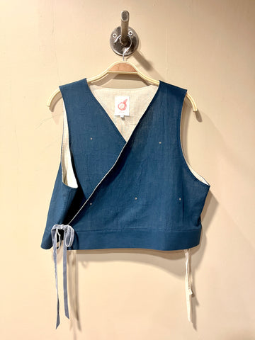 A sleeveless wrap-style vest in indigo blue fabric with white ties and white lining, hanging on a wooden hanger against a beige wall.