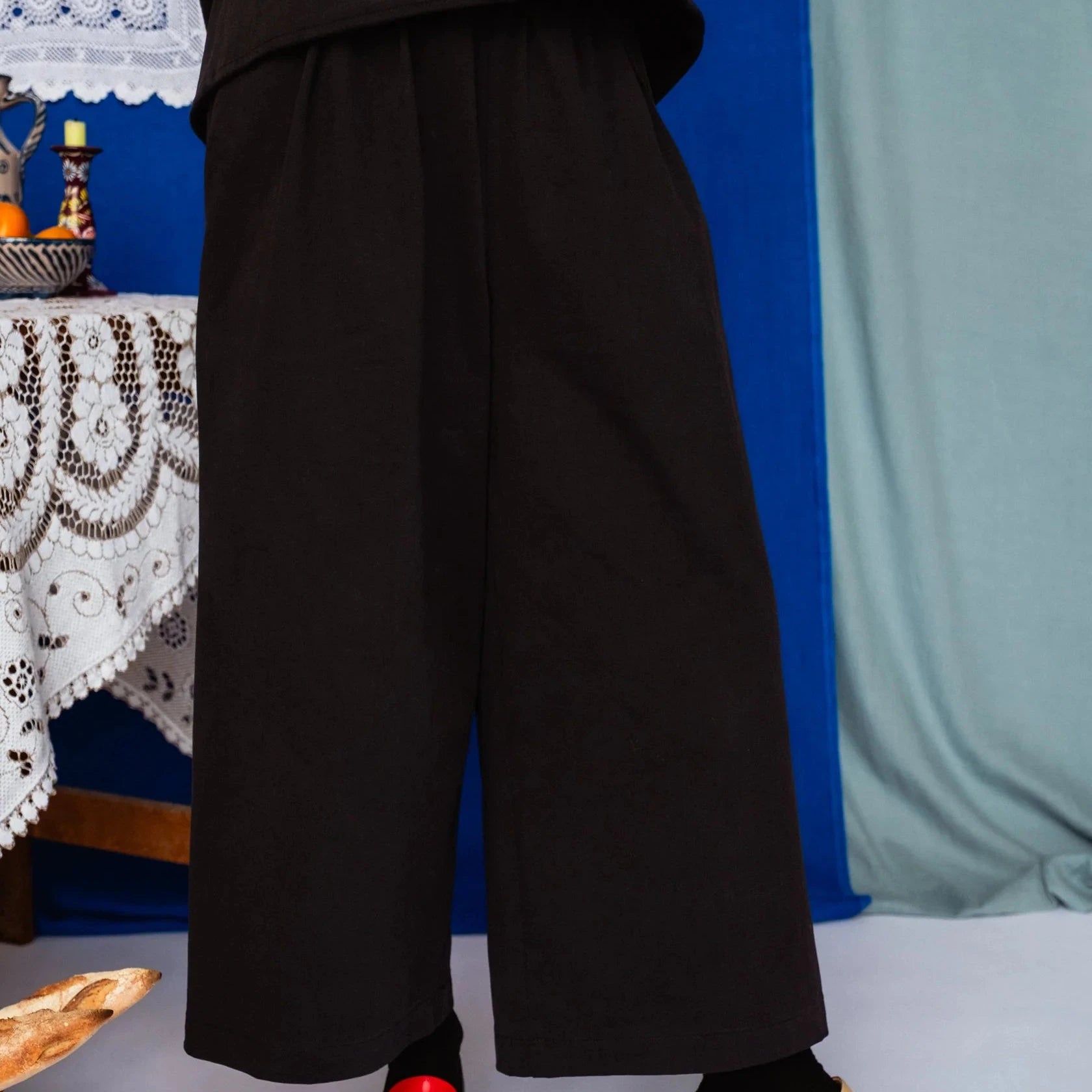 Person wearing a black outfit with wide-leg pants and sandals in a room with a blue wall and table.