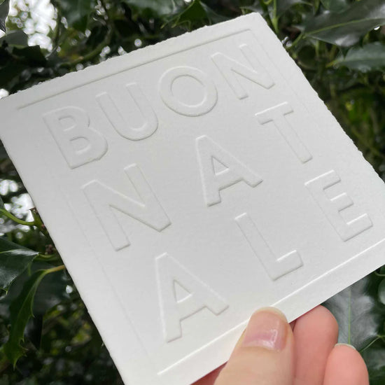 White card with 'BUON NATALE' embossed on it held against a green leafy background