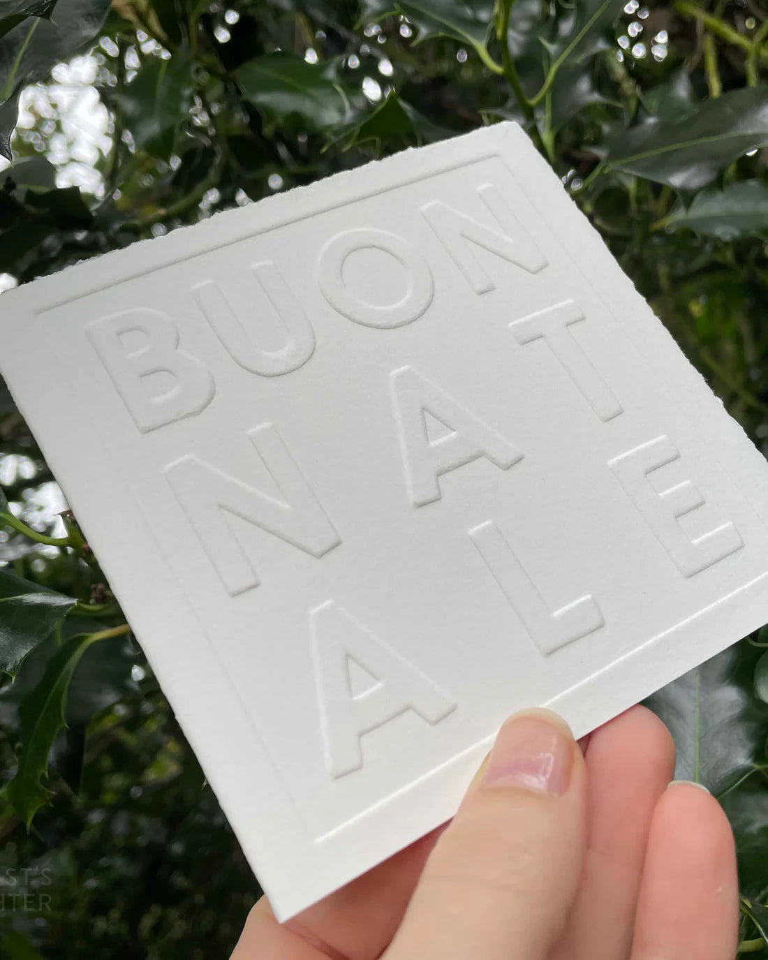 White card with 'BUON NATALE' embossed on it held against a green leafy background