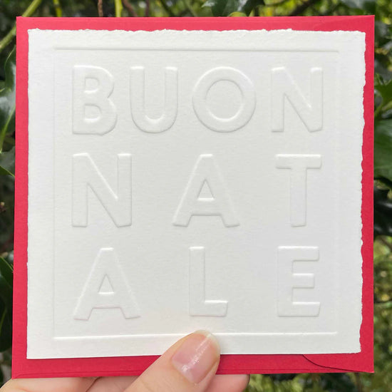 White card with 'BUON NATALE' text held against a green leafy background
