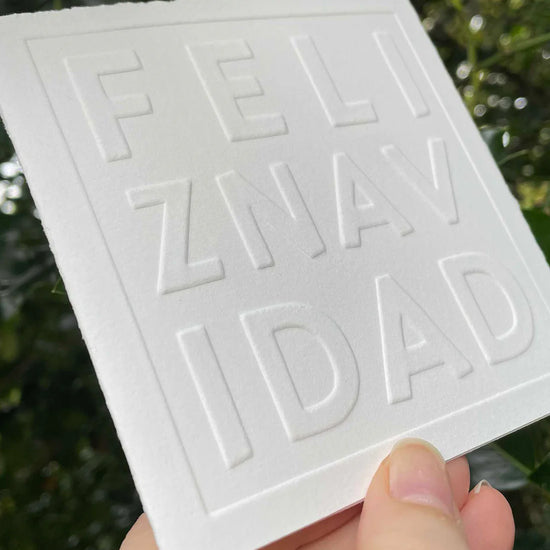 White card with embossed text 'Feliz Navidad' held against a green leafy background