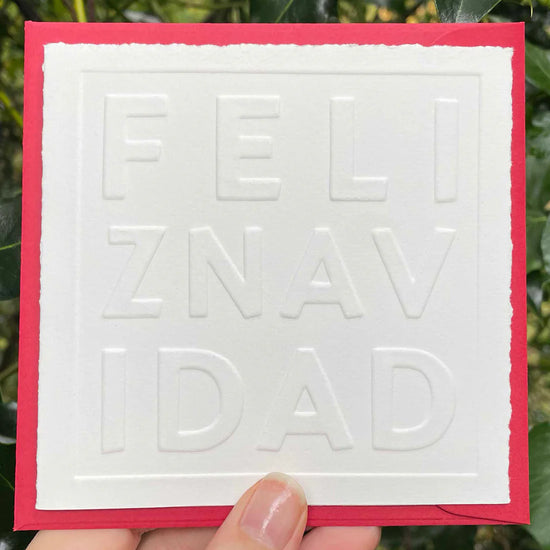 Card with embossed text 'FELIZ NAVIDAD' held against a green leafy background