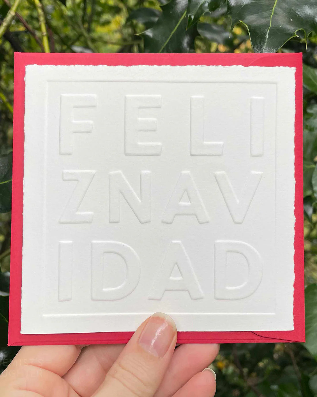 Card with embossed text 'FELIZ NAVIDAD' held against a green leafy background