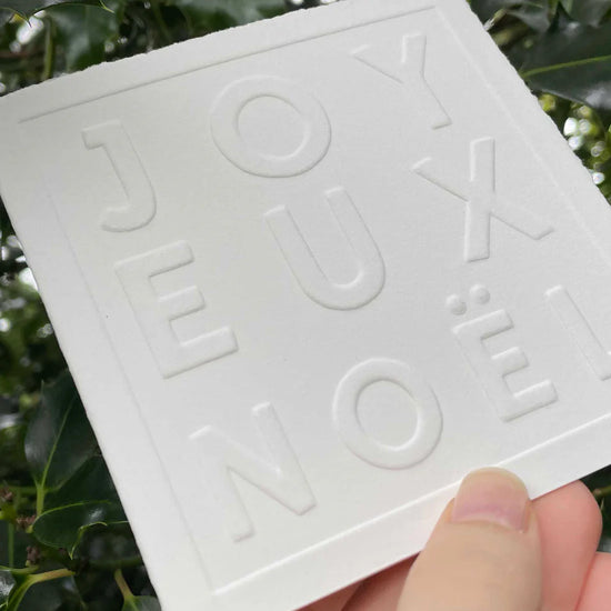 White Christmas card with 'JOYEUX NOEL' embossed text held against a green leafy background