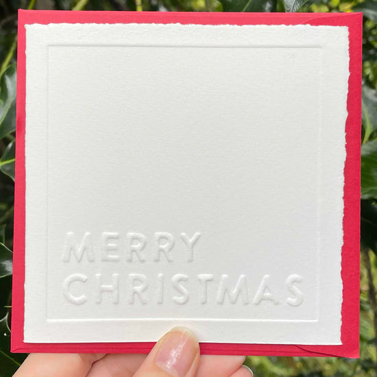 White card with 'Merry Christmas' text held against a green leafy background