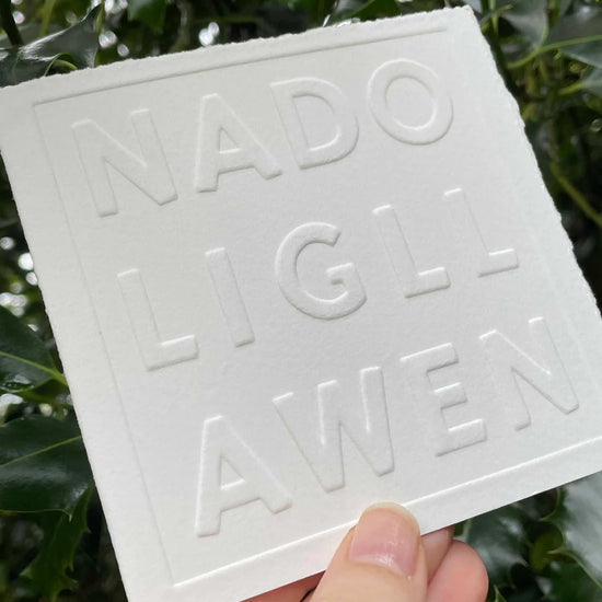 White embossed card with Nadolig Llawen text held in front of green foliage