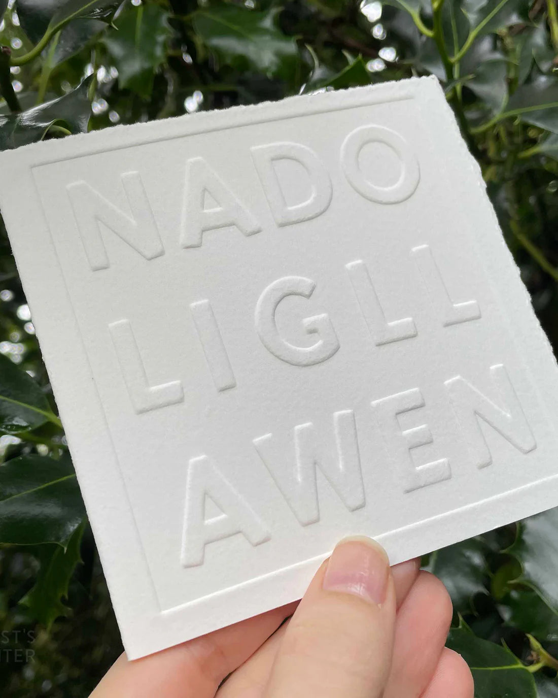 White embossed card with Nadolig Llawen text held in front of green foliage