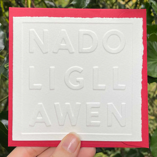 White card with embossed text 'NADOLIG LLAWEN' held against a green leafy background