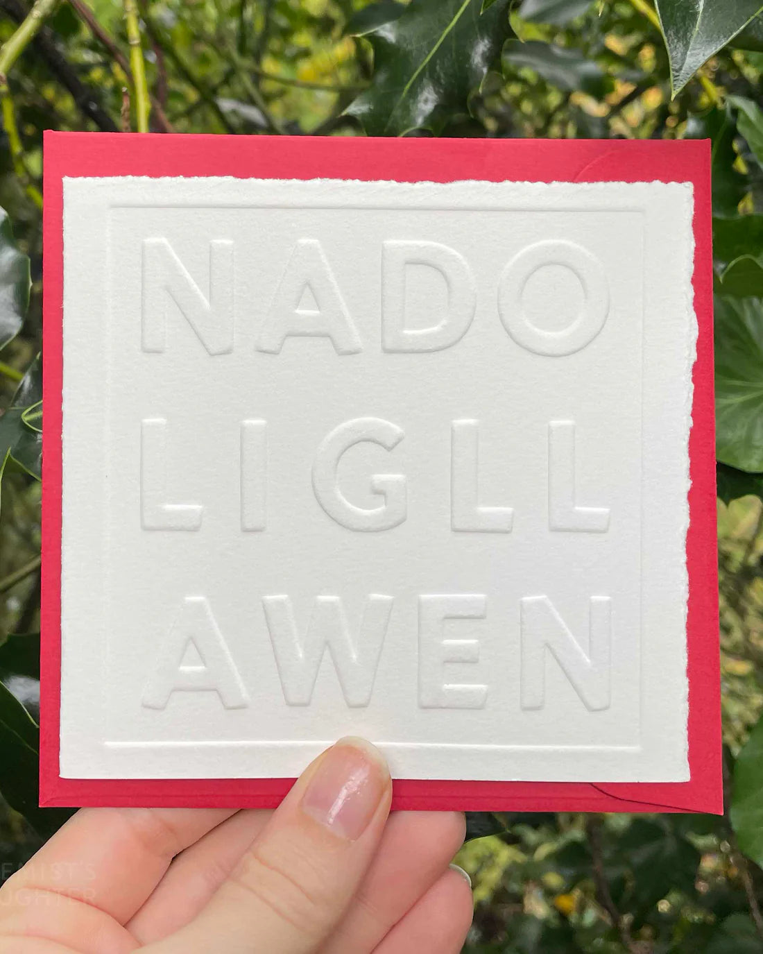White card with embossed text 'NADOLIG LLAWEN' held against a green leafy background
