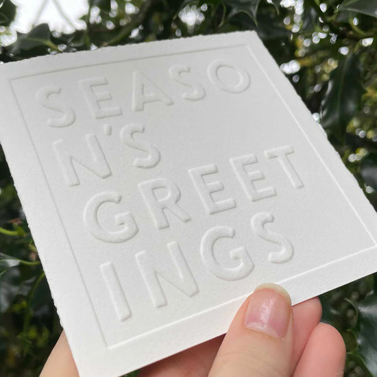 White card with 'Season's Greetings' text held in front of green foliage