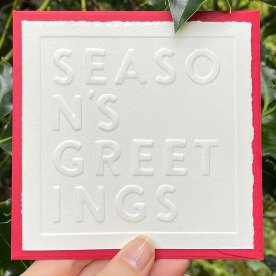 Card with 'Season's Greetings' embossed text held by a hand against a green leafy background
