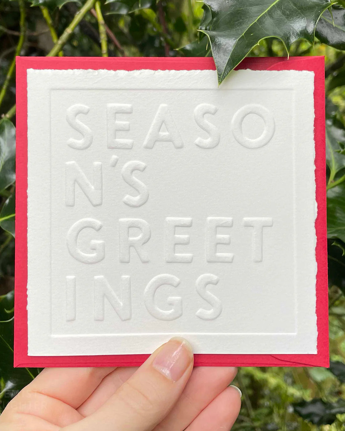 Card with 'Season's Greetings' embossed text held by a hand against a green leafy background
