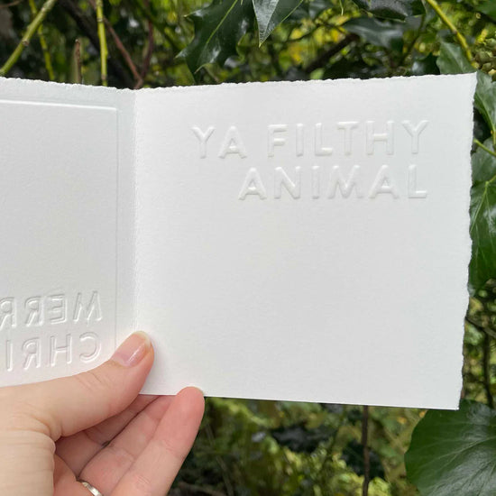 Hand holding a white card with embossed ‘YA FILTHY ANINAL’ text against a green leafy background