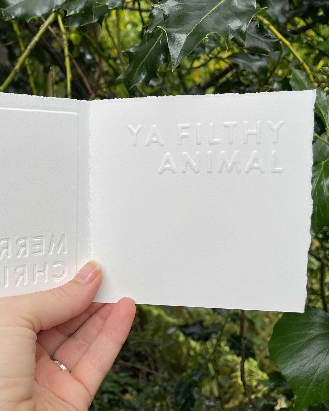 Hand holding a white card with embossed ‘YA FILTHY ANINAL’ text against a green leafy background