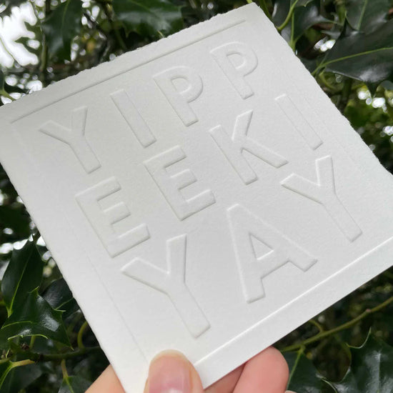White square card with 'YIPPEE KI YAY' embossed text held in front of green leaves
