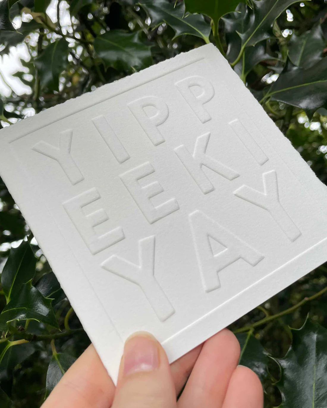 White square card with 'YIPPEE KI YAY' embossed text held in front of green leaves