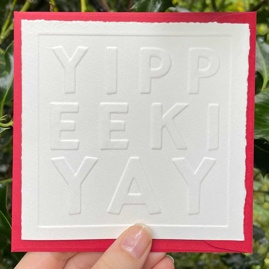 Card with embossed 'YIPPEEKIYAY' text held by a hand against a green leafy background