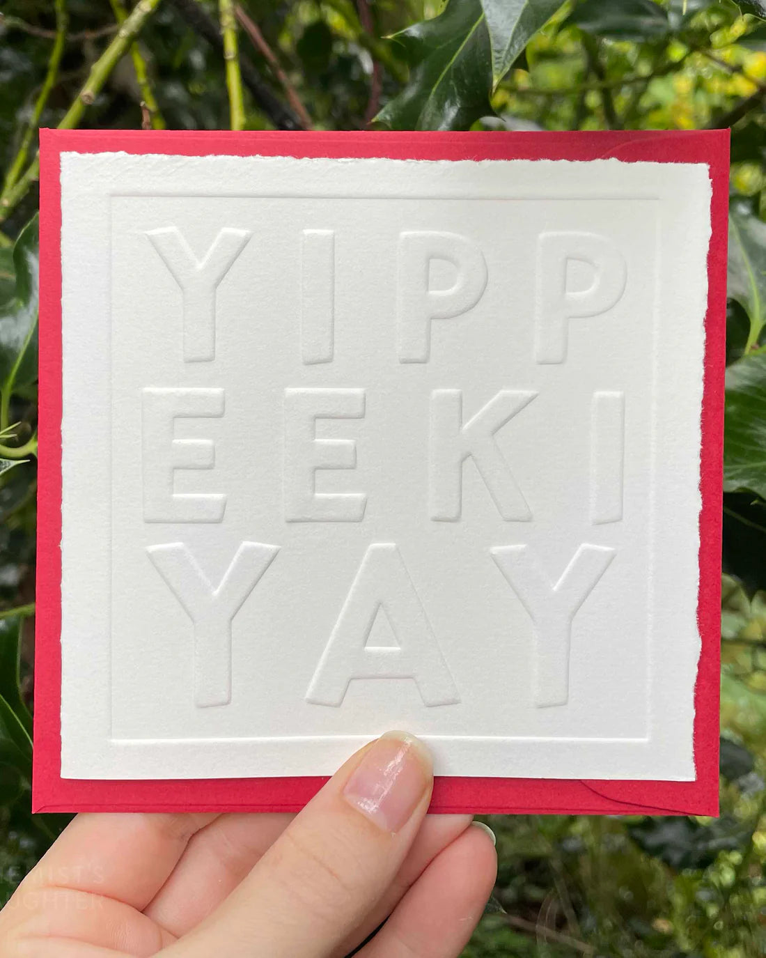 Card with embossed 'YIPPEEKIYAY' text held by a hand against a green leafy background