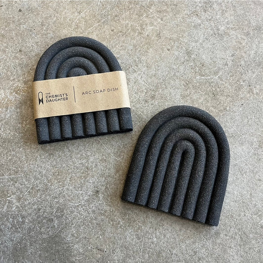 A large arch-shaped soap dish with a textured, stone-like finish, placed on a flat surface. It is black in color and has a branded tag indicating 'The Chemist's Daughter'.