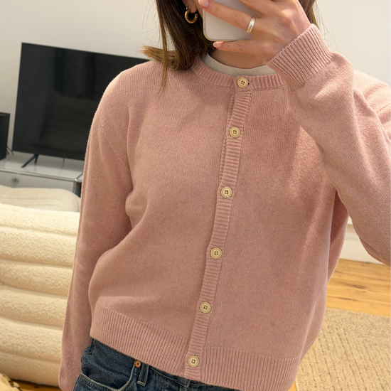 woman in blush pink lambswool knitted cardigan with cream buttons matched with jeans and gold hoops with medium length brown hair. She stands in front of a television in her living room
