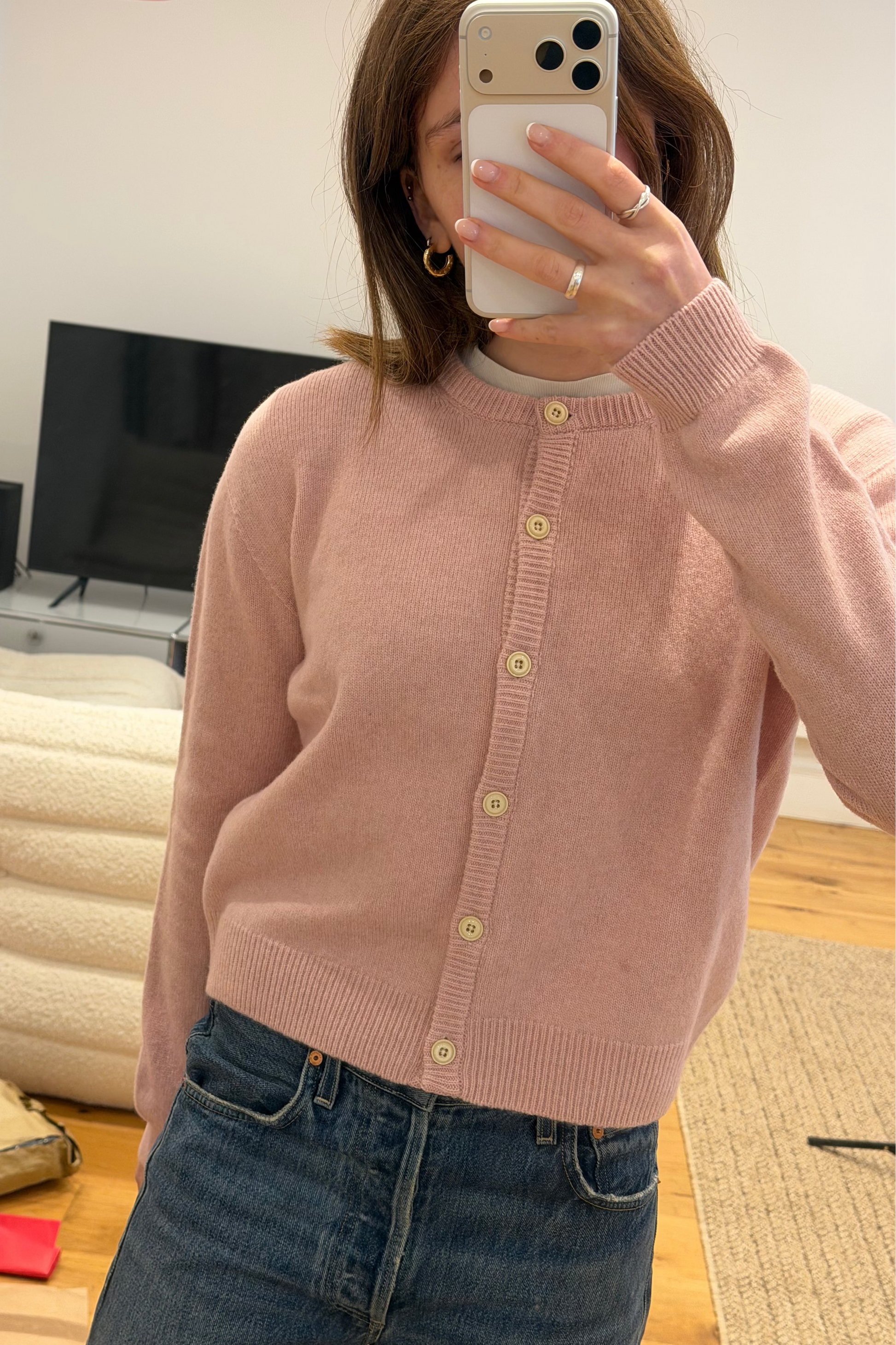 woman in blush pink lambswool knitted cardigan with cream buttons matched with jeans and gold hoops with medium length brown hair. She stands in front of a television in her living room
