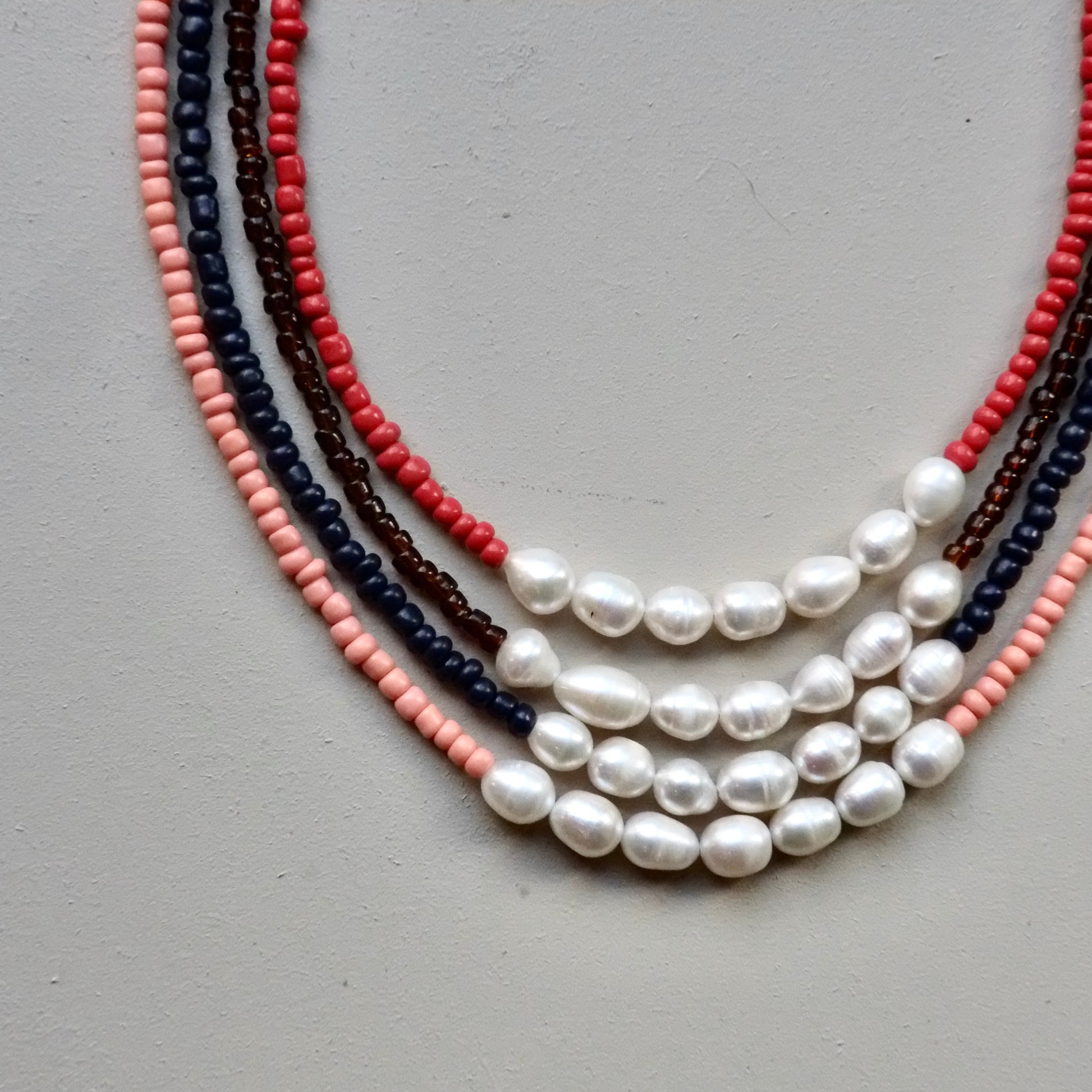 A multi-strand beaded necklace laid flat on a neutral gray background. The necklace features strands of white baroque pearls, coral red beads, dark brown beads, and navy blue beads.
