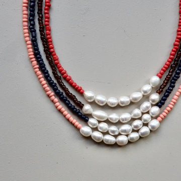 A multi-strand beaded necklace laid flat on a neutral gray background. The necklace features strands of white baroque pearls, coral red beads, dark brown beads, and navy blue beads.