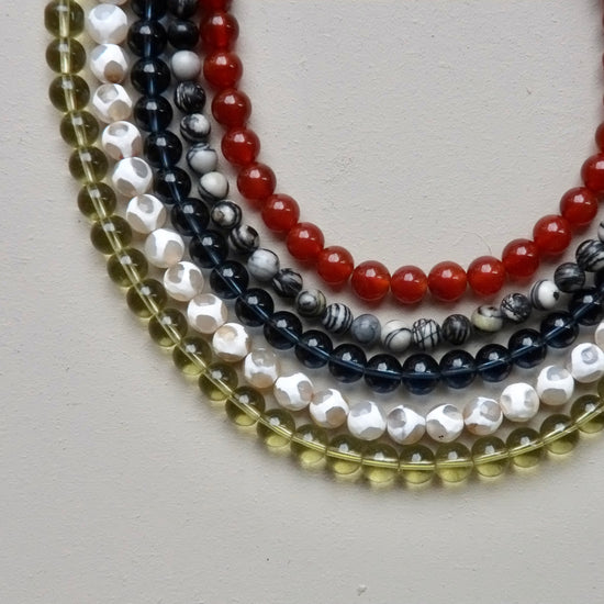 Five strands of round gemstone beads arranged in overlapping curves on a light neutral background. The beads include translucent olive green, white with black marbling, dark navy with black marbling, glossy red, and glossy white with black marbling.