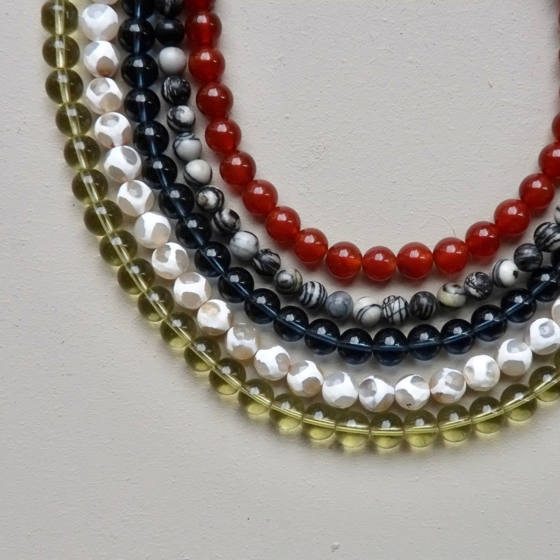 Five strands of round gemstone beads arranged in overlapping curves on a light neutral background. The beads include translucent olive green, white with black marbling, dark navy with black marbling, glossy red, and glossy white with black marbling.