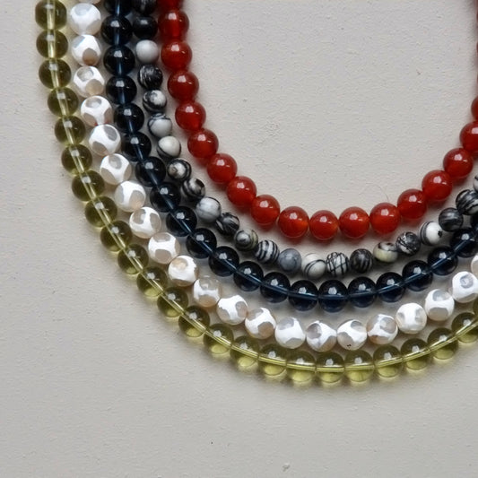 Five strands of round gemstone beads arranged in overlapping curves on a light neutral background. The beads include translucent olive green, white with black marbling, dark navy with black marbling, glossy red, and glossy white with black marbling.