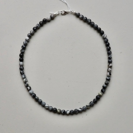 A beaded necklace made of small marbled beads in black, gray, and white, with a silver clasp, laid flat on a light gray background.