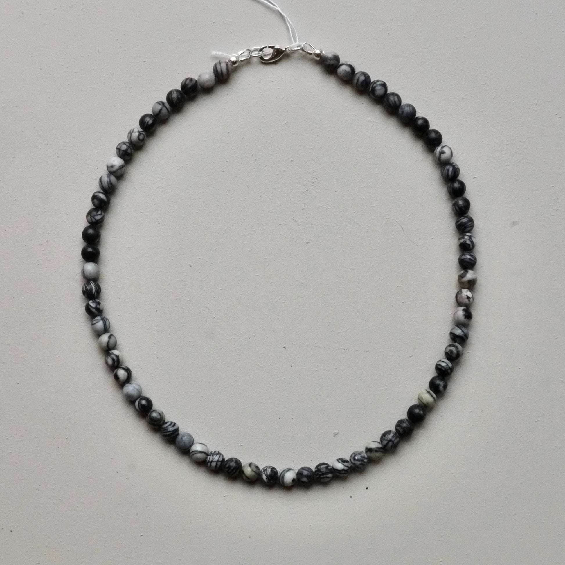 A beaded necklace made of small marbled beads in black, gray, and white, with a silver clasp, laid flat on a light gray background.