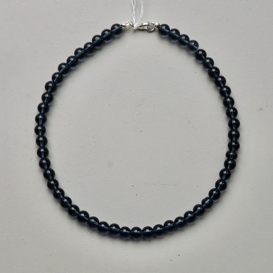 A beaded necklace made of small, dark round beads with a silver clasp, displayed on a light gray surface.