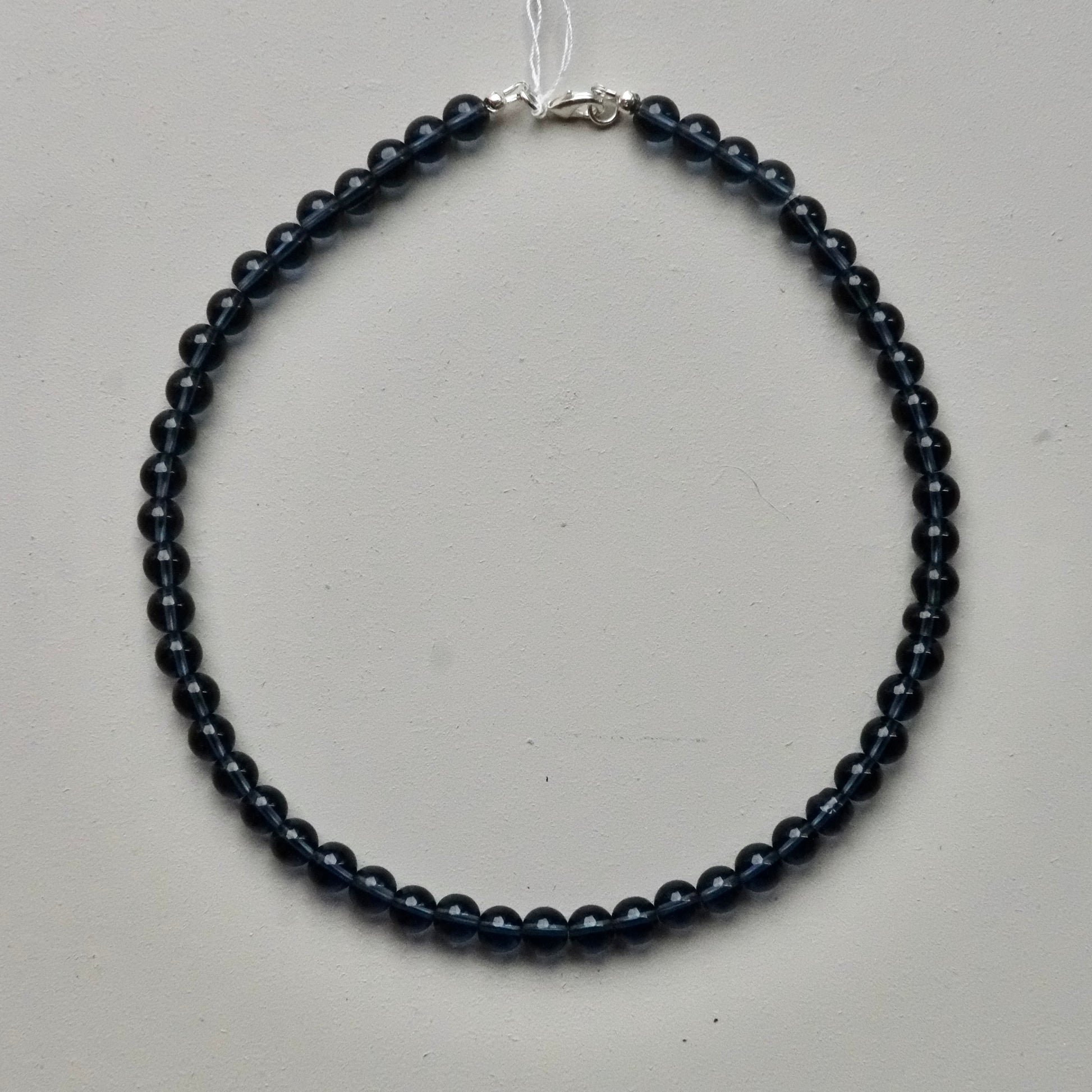 A beaded necklace made of small, dark round beads with a silver clasp, displayed on a light gray surface.