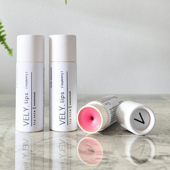 Two white VELY. lip balm tubes stand upright on a reflective surface, with one tube lying on its side showing pink balm inside.