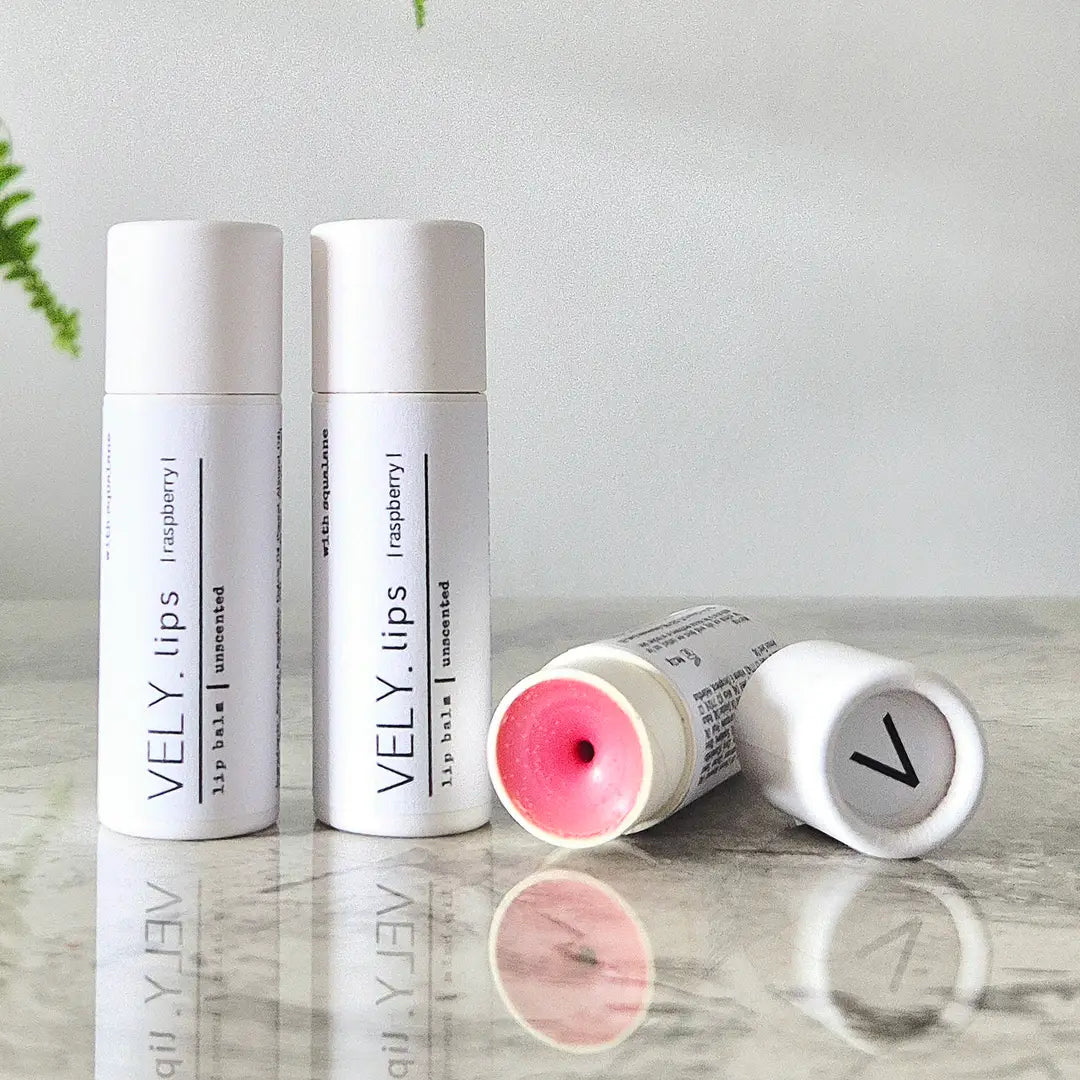 Two white VELY. lip balm tubes stand upright on a reflective surface, with one tube lying on its side showing pink balm inside.