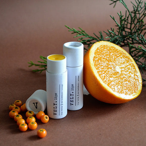 Natural lip balm in white cylindrical containers with orange slices and berries on a brown background