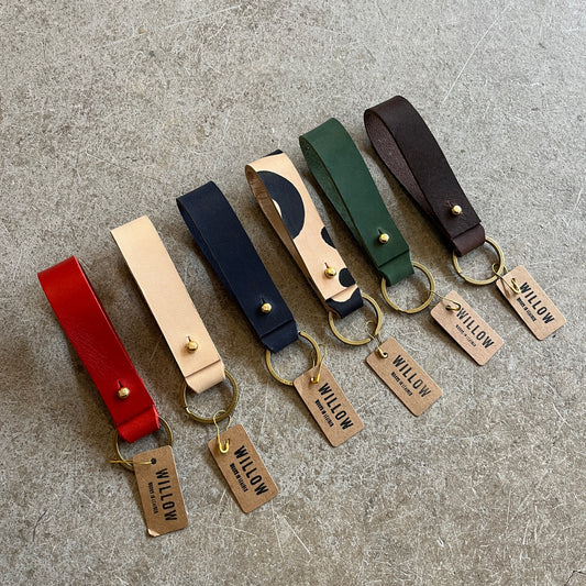 A collection of leather loop keyrings in various colors including red, navy, green, brown, and beige, with solid brass details and branded tags.