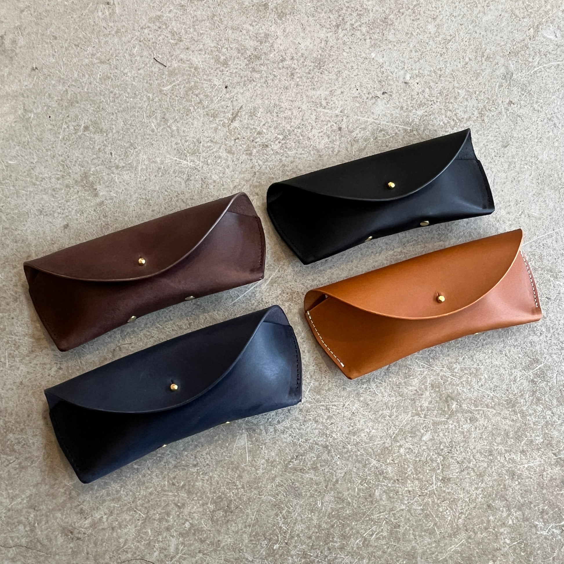 Four leather sunglasses cases in different colors (black, brown, tan) laid out on a textured surface.