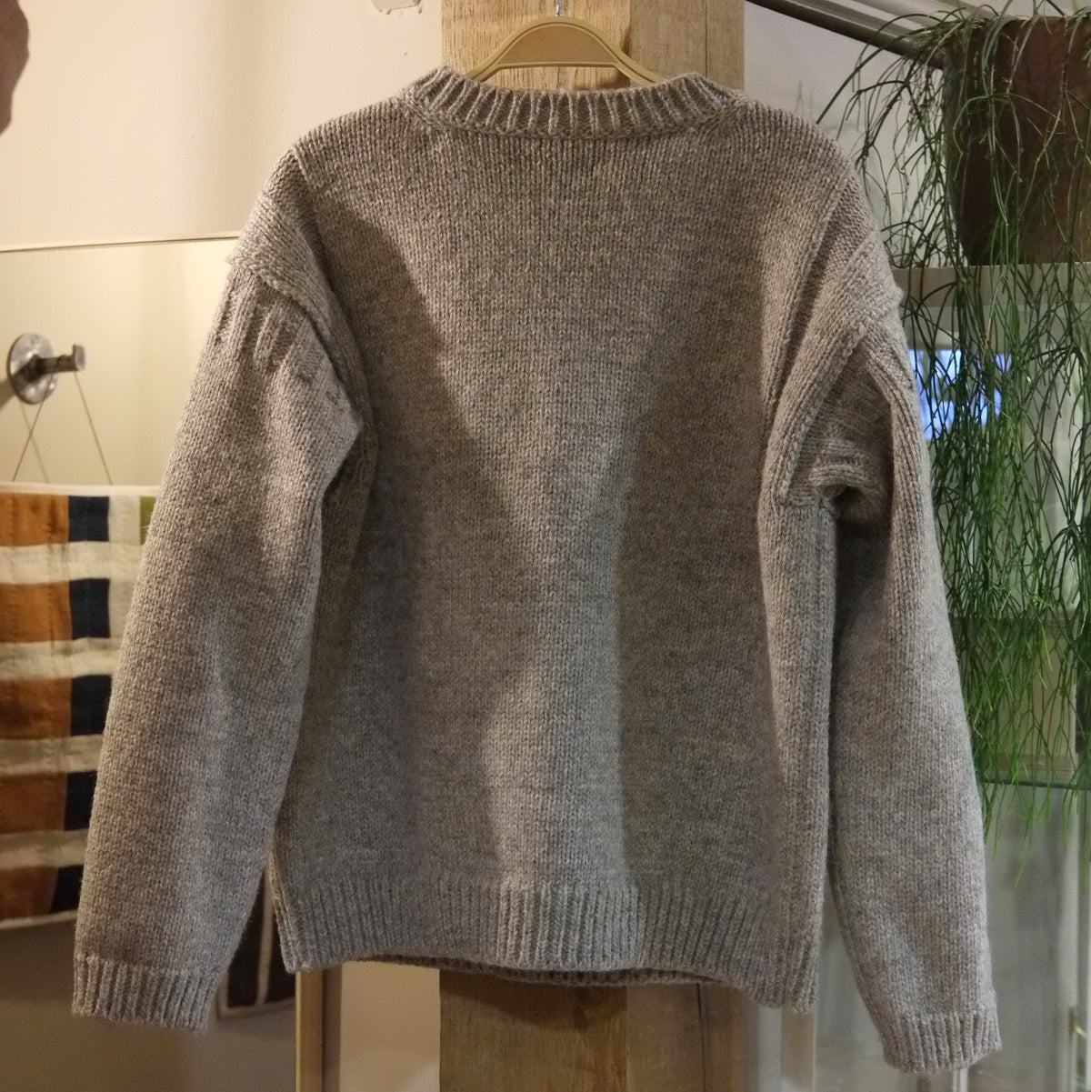 A light gray knit sweater hanging on a wooden hanger from a clothing rack inside a boutique, with rustic wooden pillars and a glass display case in the background.