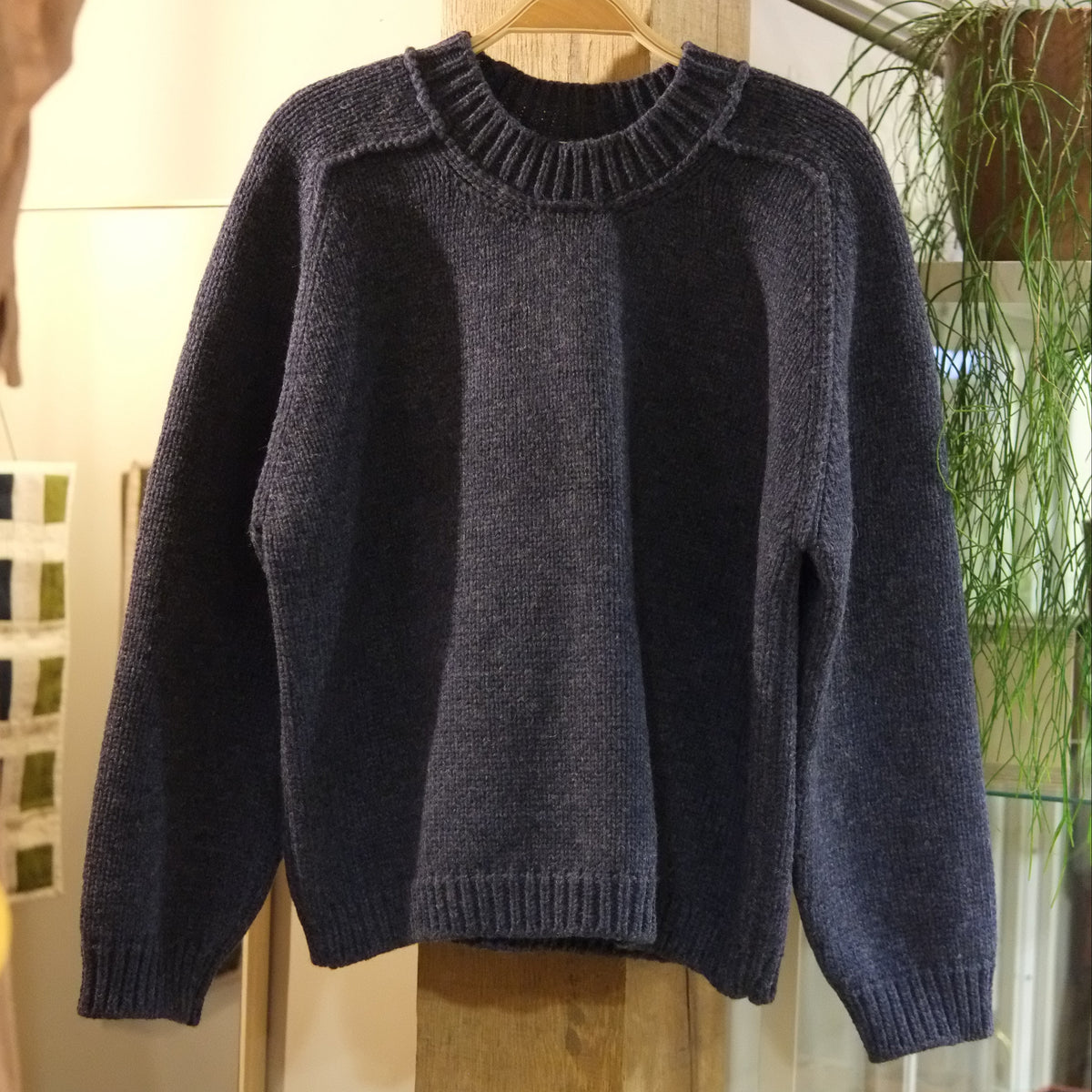 A dark navy knit sweater with a crew neck and ribbed cuffs and hem, hanging on a wooden hanger on a clothing rack inside a store.