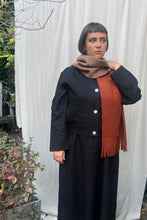Load image into Gallery viewer, Person wearing a black coat with a brown scarf and orange sweater against a white curtain background.
