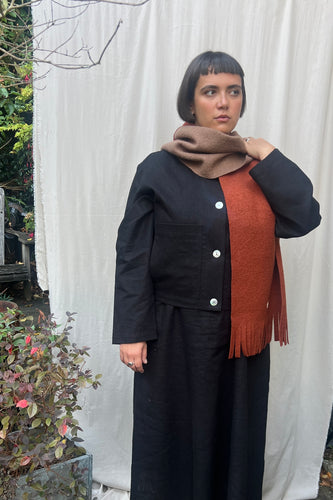 Person wearing a black coat with a brown scarf and orange sweater against a white curtain background.