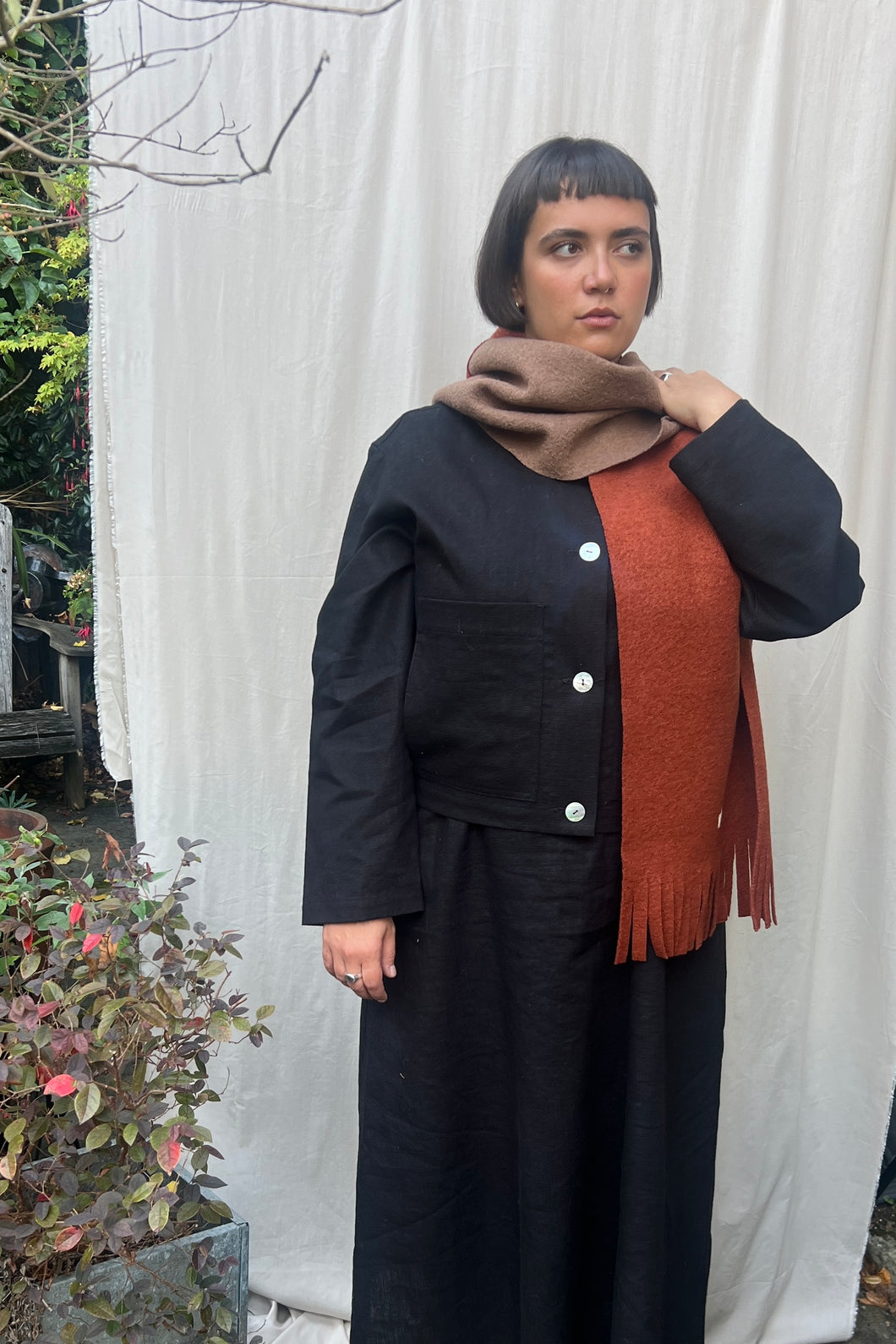 Person wearing a black coat with a brown scarf and orange sweater against a white curtain background.