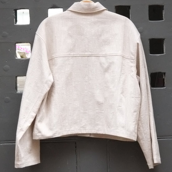 reverse of cropped jacket in a natural colour fabric photographed on a grey door