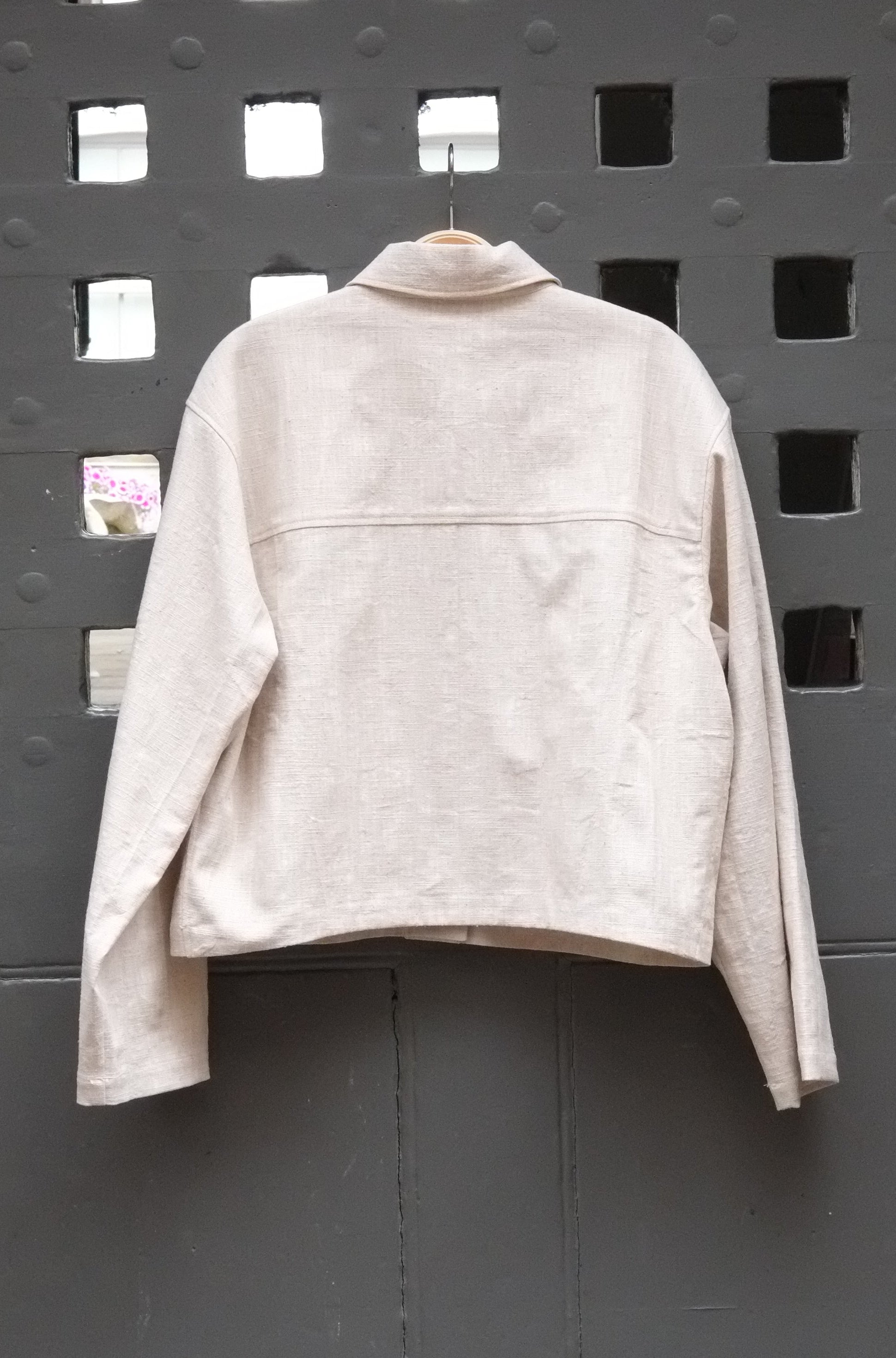 reverse of cropped jacket in a natural colour fabric photographed on a grey door