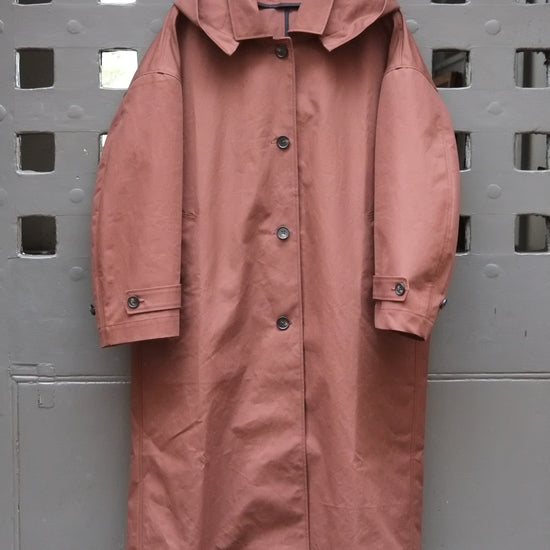 A chocolate brown hooded raincoat hanging on a wooden hanger against a grey door with a square grid pattern.