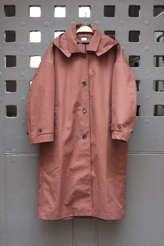 A chocolate brown hooded raincoat hanging on a wooden hanger against a grey door with a square grid pattern.