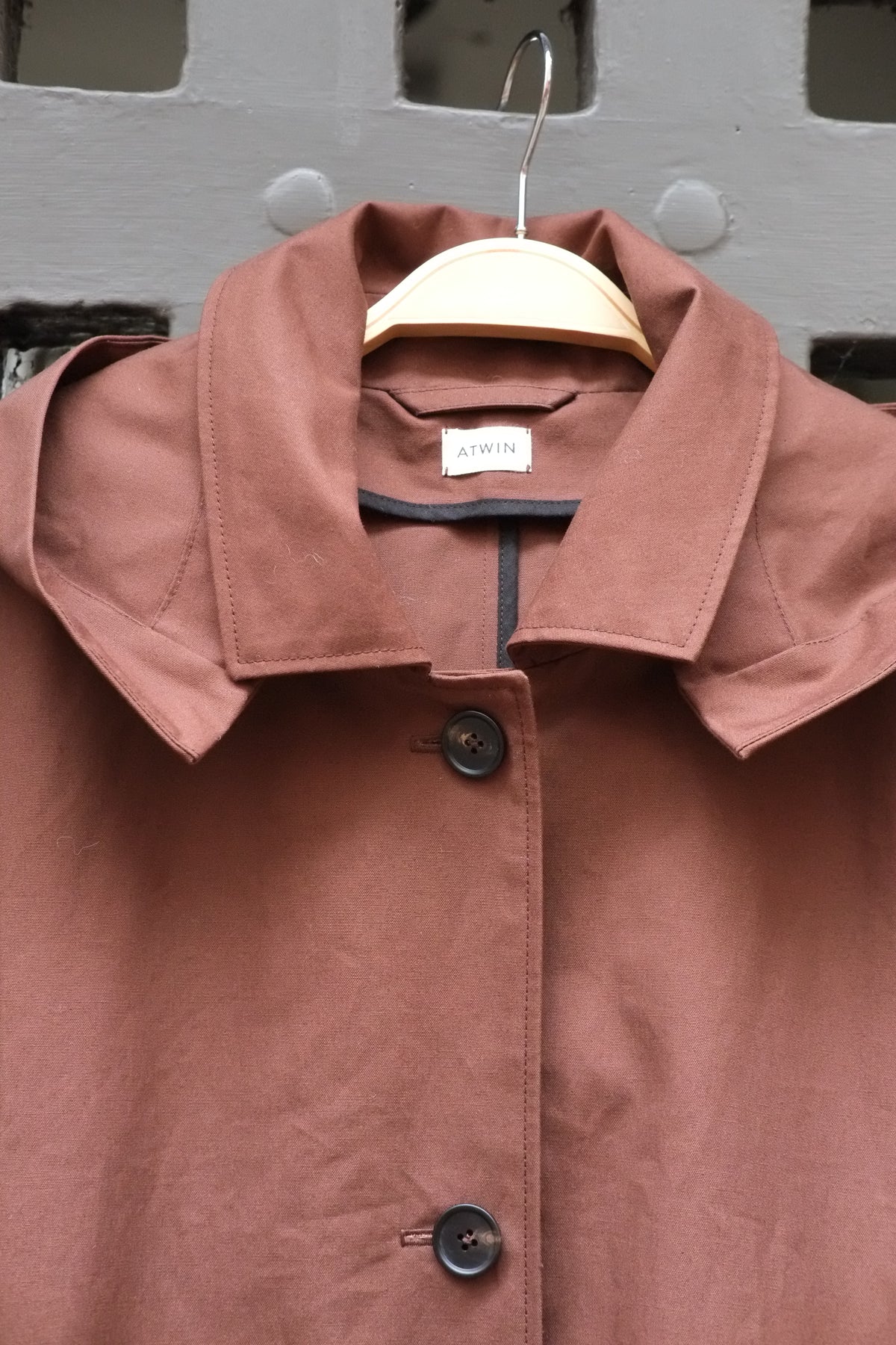 A chocolate brown  button-up jacket with a wide collar, hanging on a wooden hanger against a grey door.